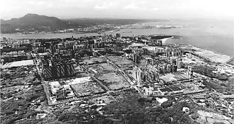 2023年臺灣某河口的黑白空拍照片，遠景為連綿山丘與海岸線，中景可見大片正在興建中的高樓建築群（多棟建築物外觀顯示尚未完工的鷹架與裸露結構），建築群周圍夾雜農地、樹林與空地，畫面右側緊鄰海岸與沙灘，左側為原有市區聚落