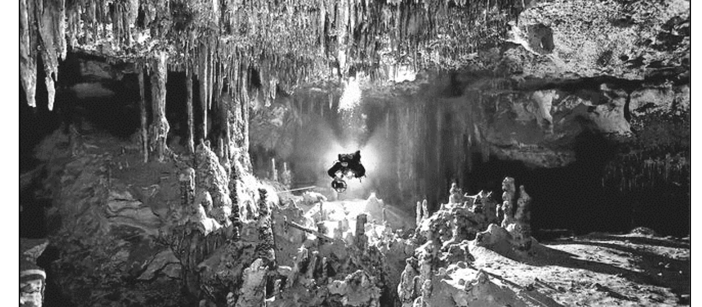 照片1：黑白照片，猶加敦半島被地下水淹沒的石灰岩洞穴（cenote），畫面上方懸垂大量鐘乳石與石筍，下方水面倒映洞頂景象，一名配戴頭燈、攜帶氣瓶的潛水人懸浮在畫面正中央的水中，四周環繞著自水底聳立的石筍地形，洞穴左右兩側可見更深邃的黑暗通道向外延伸，顯示洞穴彼此相連的特徵